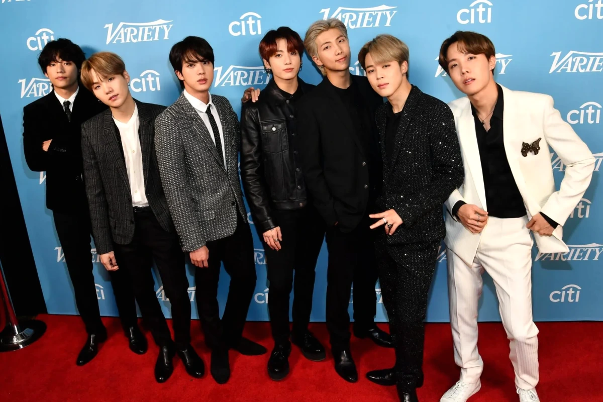 BTS sur scène sous des projecteurs violets, avec le nom « Drake » affiché en arrière-plan sur un écran