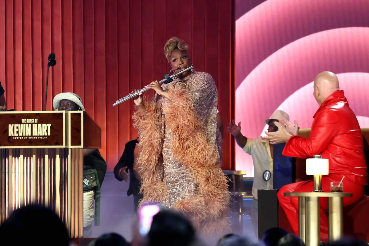 Lizzo sur scène tenant une flûte, souriante, lors du roast de Kevin Hart, micro à la main.